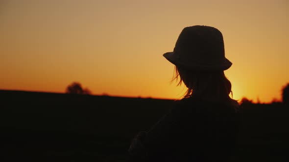 A Young Woman in a Hat Admires the Sunset in a Picturesque Place alt