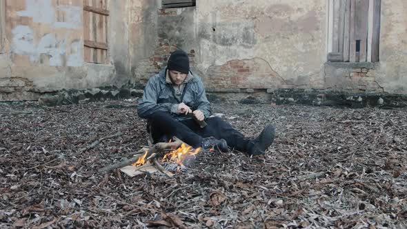 Homeless Man Sits on Ground and Pushes Pieces of Cardboard Into Fire to Keep Warm From Cold alt