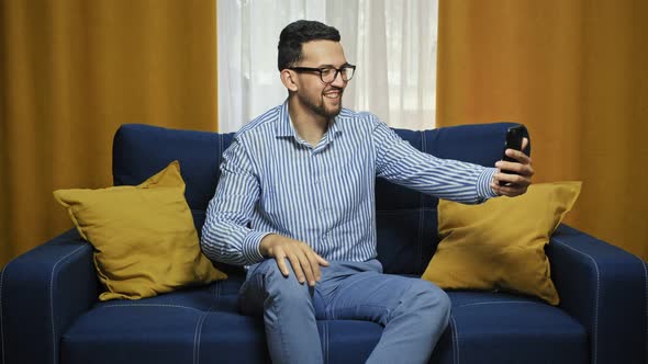 Smiling Man in Shirt with Smartphone Making Video Call While Sitting on Couch in Living Room alt