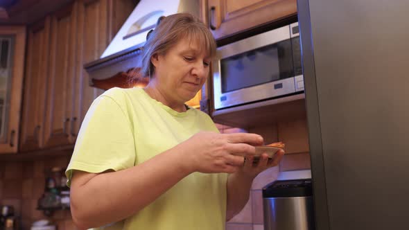 Hungry middle aged woman opens a refrigerator at night and eating hamburger with sausage.