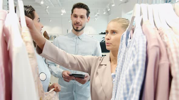 Young Women Friends Choosing Clothes and Talking to Salesman Holding Tablet in Shop alt
