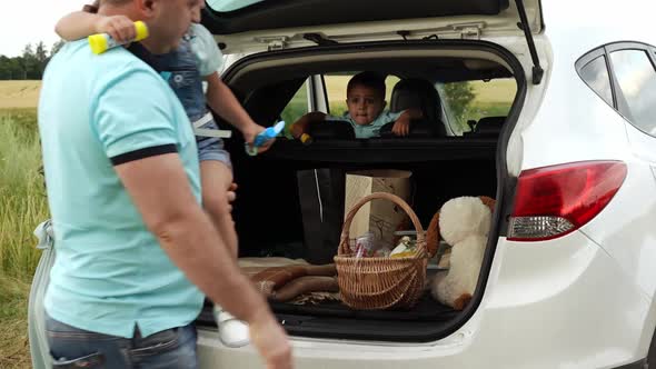 Couple with Kids Arrived at Picnic in Field By Car and Unload Luggage From Trunk in Slowmo alt