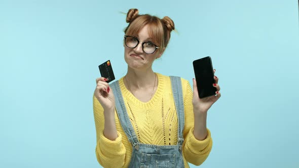 Cheerful Young Woman in Glasses and Overalls Showing Smartphone Screen and Plastic Credit Card Using