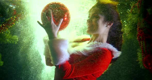 Woman Dressed in Suit Stylised Like Santa Claus Is Playing with Red Shiny Ball Underwater alt