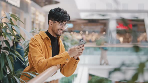 Happy Smiling Young Arab Guy Stands with Telephone in Hands Typing Message on Dating Site Chatting alt