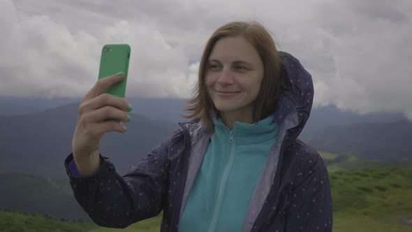 30s Woman Traveler Taking Selfie Picture on Cell Phone Standing in Cloudy Mountains alt