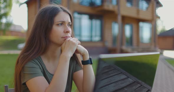 Frustrated Young Woman Sitting in Front of Laptop at Table in Courtyard of the House alt