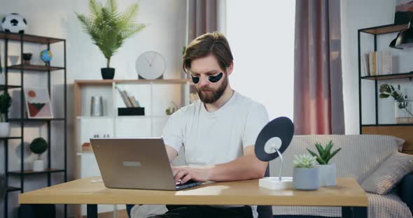 30-Aged Bearder with Anti-Aging Hydrogel Eye-Patches Sitting at the Table and Working on Computer alt