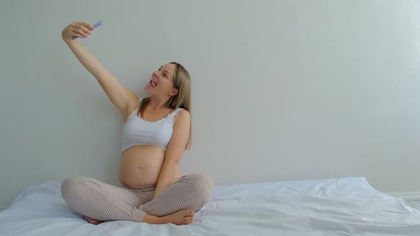 happy pregnant woman with smartphone taking selfie in bed at home