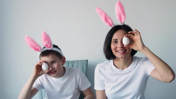 Playful boy and woman with bunny ears keeping white eggs near eyes on white background. Happy child