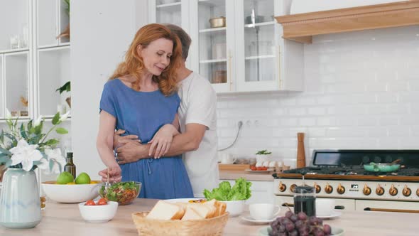 Aged Man Hugging His Attractive Wife in Kitchen alt