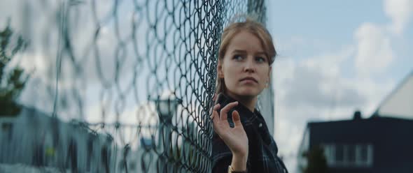 Young woman leaning towards metal fence while waiting for someone alt