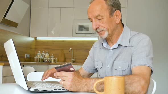 Trendy Mature Man is Working From Home with Laptop Sitting at the Table in His Kitchen Buying alt