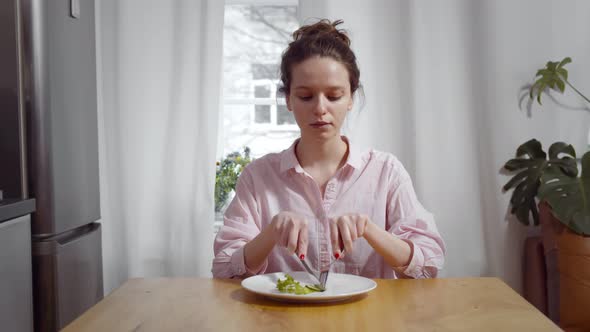 Young Skinny Woman Sitting at Table and Eating Piece of Cucumber and Salad with for and Knife alt