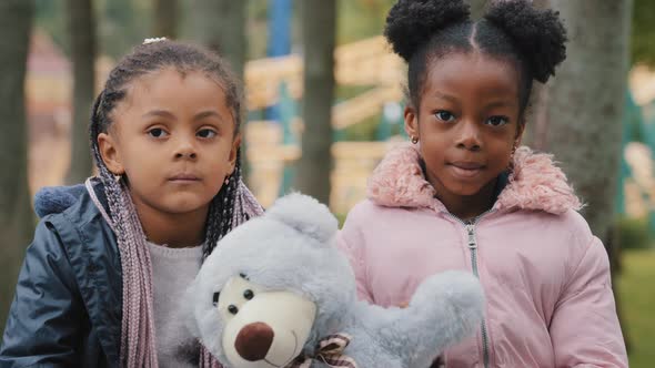 Two Friends Sitting on Bench in City Park Kid Waving Paw Teddy Bear African American Little Girls alt