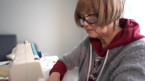 Pensioner Woman Wearing Glasses Sews Home Textiles on a Sewing Machine alt