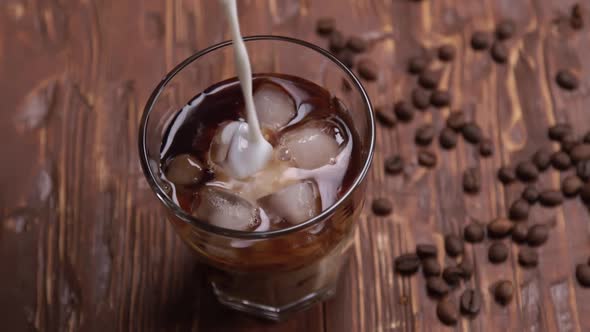 Top View Slow Motion of Cream Being Poured Into a Glass of Cold Brew Iced Coffee on Brown Wood Table