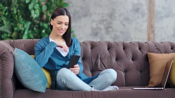 Full Shot Happy Informal European Young Woman Sitting on Couch Using Telephone alt