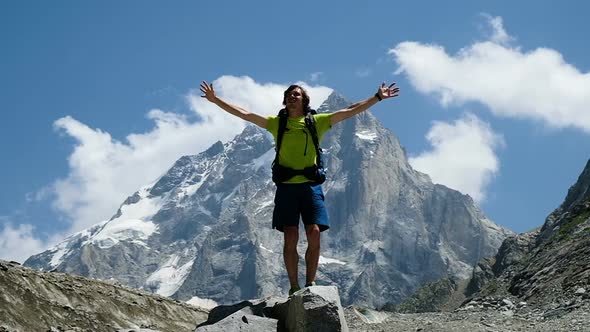 Man Tourist with a Backpack Raises His Hands Up in the Mountains in the Campaign After Climbing alt