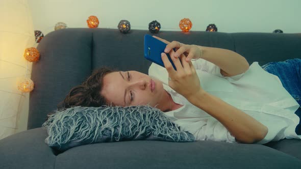 Young Woman is Using Smart Phone and Lying on Couch Leisurely in Living Room alt