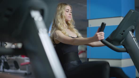 Blond Concentrated Caucasian Sportswoman Riding Exercise Bike in Gym Indoors alt
