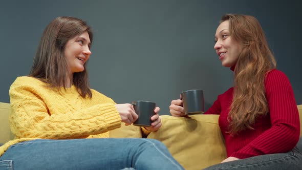 Two Smiling Girls Discuss Gossip Sitting on Cozy Yellow Sofa Drinking Coffee