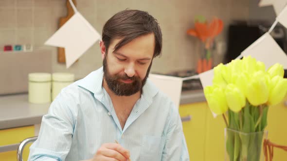 Millennial man painting Easter egg at dining table. Medium shot