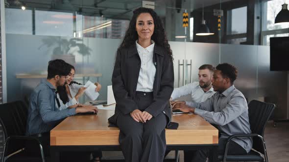 Satisfied Smiling Young Hispanic Businesswoman Sitting Carelessly on Table in Boardroom Multiracial alt