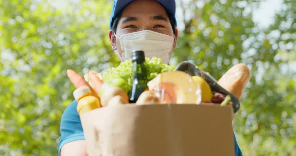 Smiling delivery man in blue uniform wearing face mask giving a grocery bag to the customer