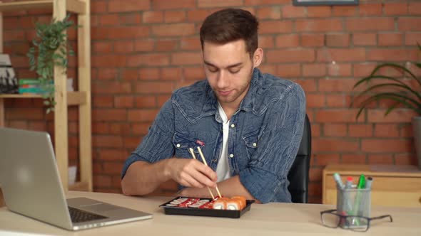 Handsome Male Freelancer Eating Sushi Rolls While Reading News on Laptop in Desk at Home Office alt