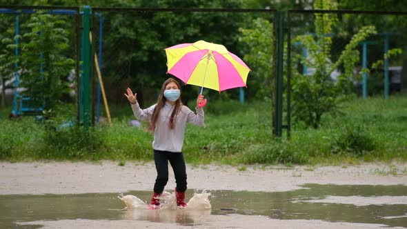 Little girl with an umbrella in a medical mask and rubber boots jumps through puddles on a spring 