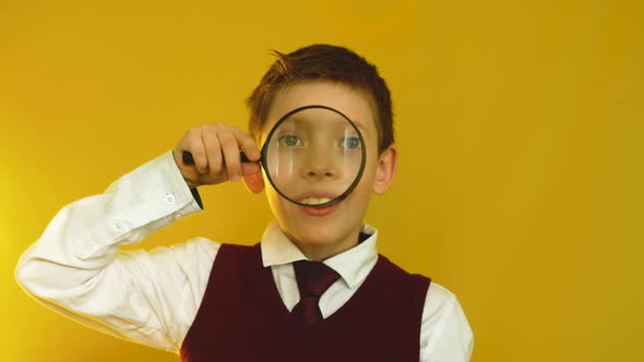 Caucasian schoolboy of 7-9 years old in a school uniform looks through a magnifying glass alt