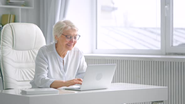 Smiling aged lady with grey hair and glasses types on laptop alt