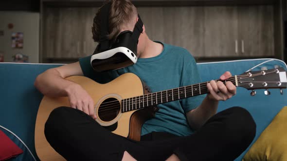 Young Man Sitting on Sofa with the Virtual Reality Glasses Is Playing at the Guitar alt