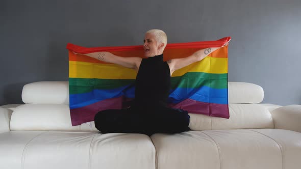Lesbian having fun and waving LGBT flag at home while sitting on white sofa