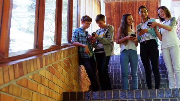 Group of smiling school friends using mobile phone in corridor alt