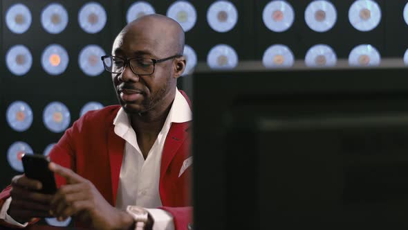 Black Bald Bearded Businessman in Red Suit and Glasses Looks at His Smartphone