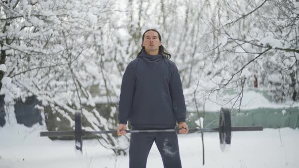 a Man Athlete with Long Hair Trains Biceps in the Winter in the Snow Raises a Barbell alt