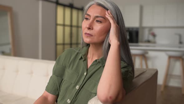 Worried Grayhaired Woman Holding Forehead with Hand Sitting on the Sofa at Home
