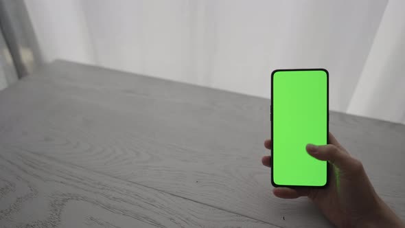 Man Showing and Using Smarphone with Green Screen While Sitting Behind White Oak Table alt