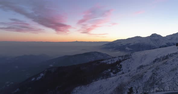 Backward Aerial Top View Over Winter Snowy Mountain and Woods Forest at Sunset or Sunrise