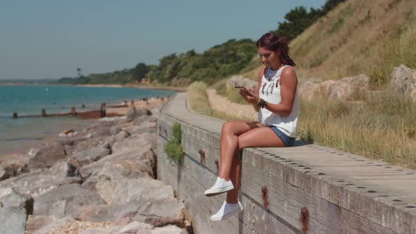 A Young Attractive Woman Sat By The Sea Using Her Phone To Order A Product Online alt