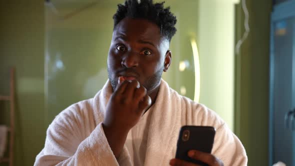 Young Handsome African American Man Brushing Teeth Watching Videos in Social Media on Smartphone alt