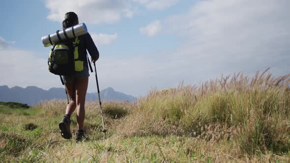 Mixed race woman wearing backpack using nordic walking poles hiking in countryside alt