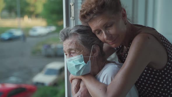 Adult Daughter Embracing with Loving Senior Mother 90 Years in Mask Standing at Window Relatives alt