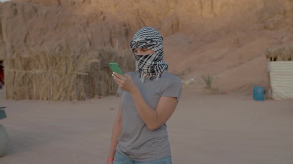 Female Tourist in Checkered Keffiyeh Standing in the Desert Using Mobile Phone alt