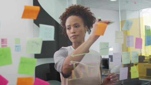 Mixed race businesswoman brainstorming, drawing with green marker on transparent board alt