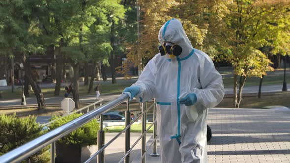 A Virologist in Protective Clothing Disinfects the Railing with an Antiseptic Spray and a Tissue alt
