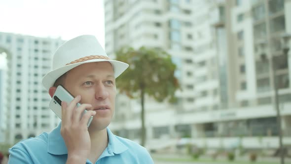 Man in a Hat Talking on a Mobile Phone While Standing on the Background of Buildings alt