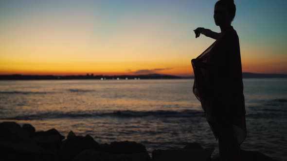 Silhouette of Woman Covered in Scarf Pointing at Sea Horizon with Dramatic Sunset Sky Above the Sea alt